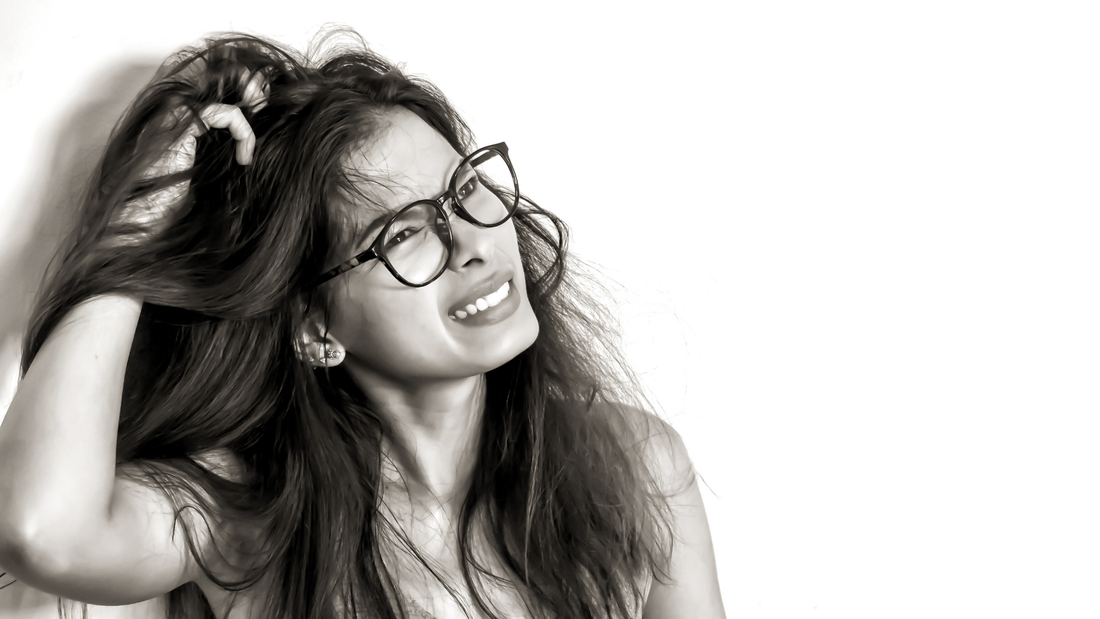 Woman with glasses showing discomfort while scratching an itchy, irritated scalp against a white background.