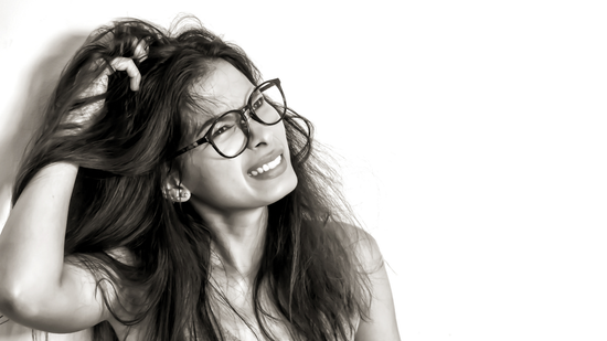 Woman with glasses showing discomfort while scratching an itchy, irritated scalp against a white background.